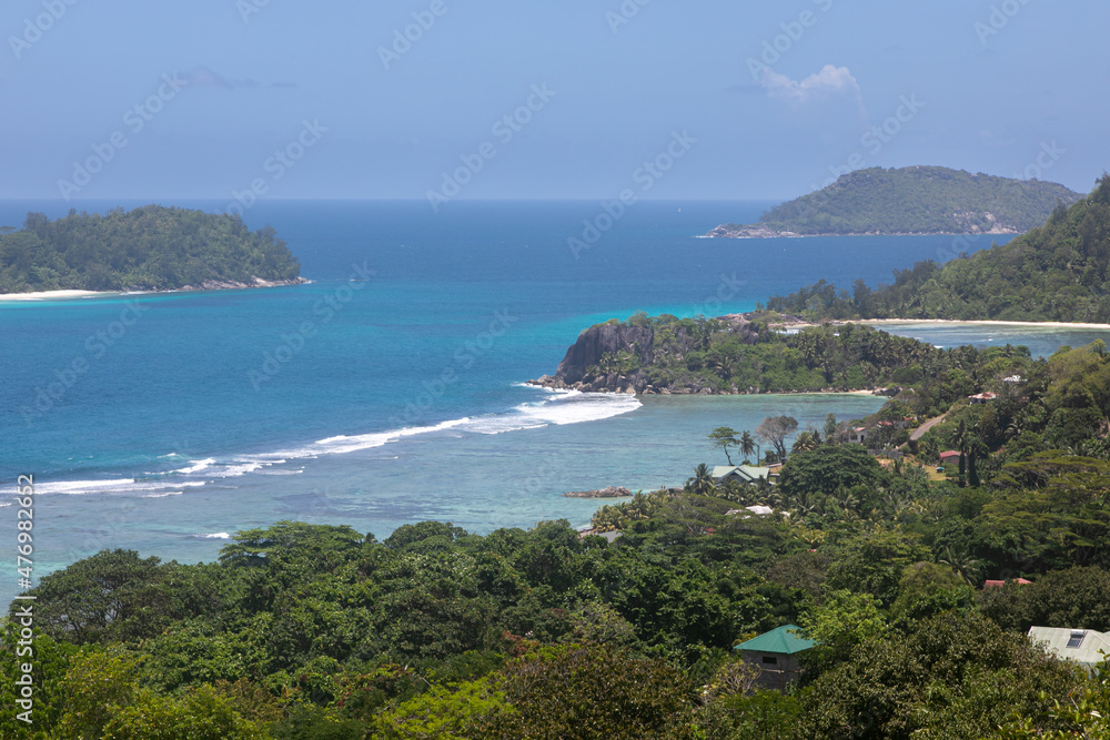 Panoramic views of the tropical island coastline. Shore is covered in white sands and green rainforest. In the distance are small islets with massive granite formations. Blue lagoon with oceanic surf.