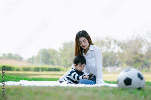 Asian mother and her little son sitting on grass and watching mobile phone in spend sunny day in green park. Happy childhood. Holiday activity and lifestyle concept.