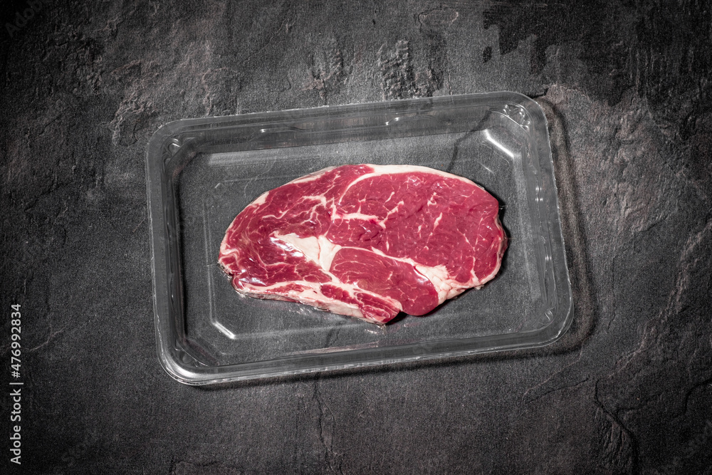 Raw beef rib steak in vacuum plastic packaging on dark stone background. Entrecote meat on black slate kitchen table. Fresh, organic, premium quality food. Cooking recipe. Copy space