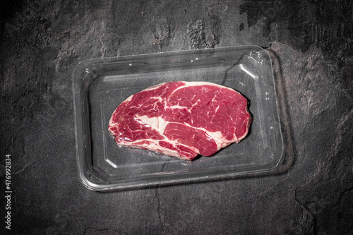 Raw beef rib steak in vacuum plastic packaging on dark stone background. Entrecote meat on black slate kitchen table. Fresh, organic, premium quality food. Cooking recipe. Copy space
