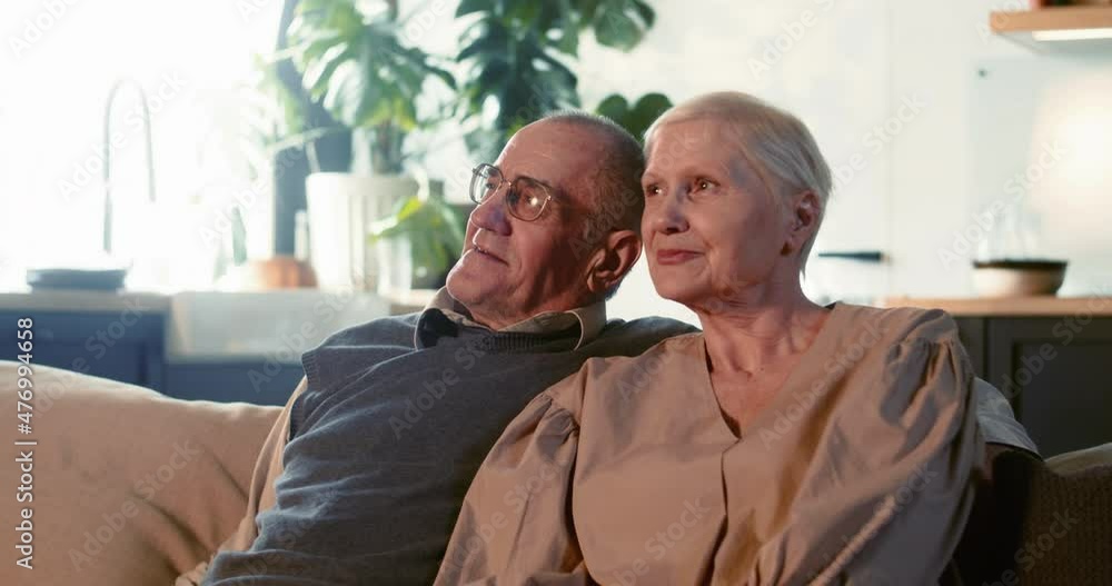 Hope concept. Portrait of happy serene retired 60s senior husband and wife smiling sitting on couch at light cozy home. Hope concept. Portrait of happy serene retired 60s senior husband and wife smiling sitting on couch at light cozy home.