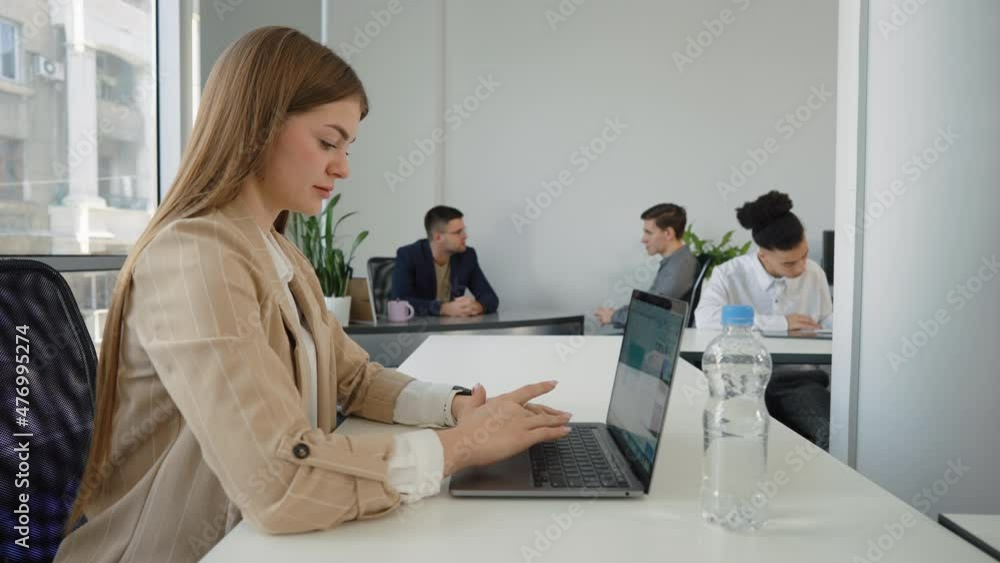 Businesswoman with long hair wearing jacket working on laptop in open space office, diverse coworkers on background. Concept of business Businesswoman with long hair wearing jacket working on laptop in open space office, diverse coworkers on background. Concept of business