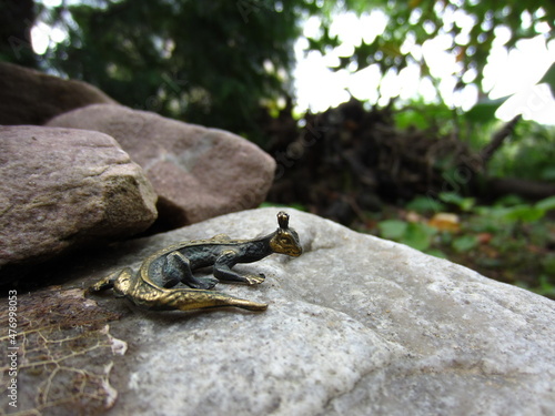 A small elegant metal lizard (garden decor) on a slide made of different stones, on the background of the garden. In the frame on the stone is an old tree leaf. © Марина Бакушева