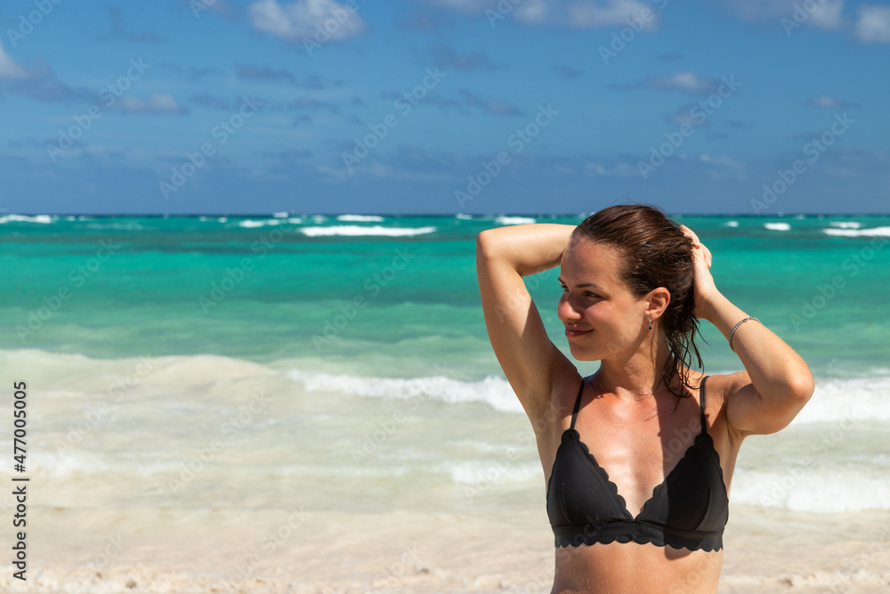 Happy attractive adult 34-years-old woman in a swimming suit stands near the ocean and enjoys the sun and her vacation in a tropical climate. Wellbeing and happiness concept. Happy attractive adult 34-years-old woman in a swimming suit stands near the ocean and enjoys the sun and her vacation in a tropical climate. Wellbeing and happiness concept.