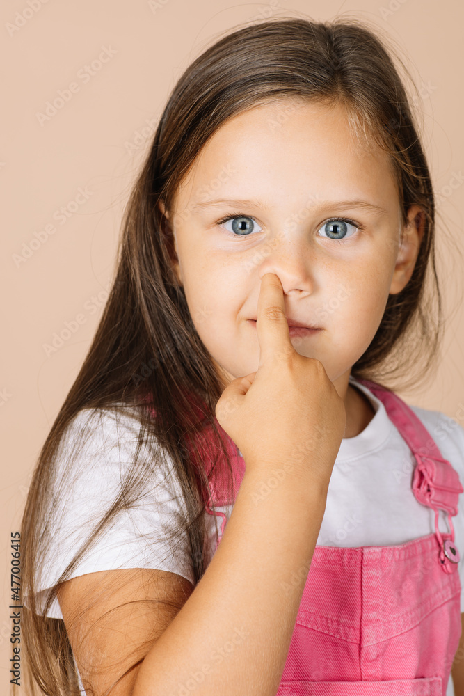 Close-up portrait kid picking nose with point finger with shining eyes smiling looking at camera wearing bright pink jumpsuit and white t-shirt on beige background. Concept of bad habits Close-up portrait kid picking nose with point finger with shining eyes smiling looking at camera wearing bright pink jumpsuit and white t-shirt on beige background. Concept of bad habits
