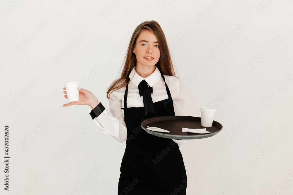 Portrait of beautiful waitress with straight hair, wearing uniform holding black round tray with takeaway coffee cup and a coffee cup in her hand on white background Portrait of beautiful waitress with straight hair, wearing uniform holding black round tray with takeaway coffee cup and a coffee cup in her hand on white background