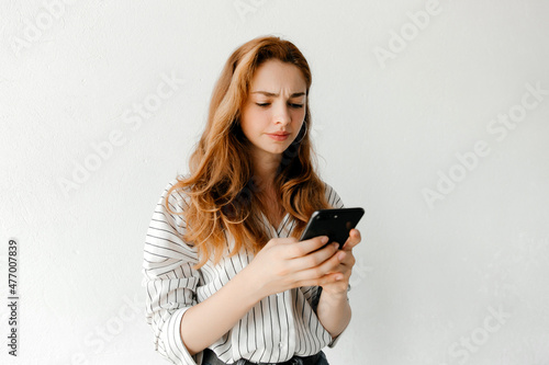 Confused young redhead woman sending a message to a group chat with the mobile, isolated over white background