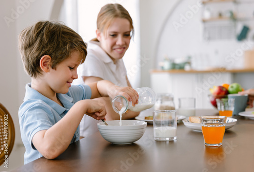Smiling little boy pouring milk into the bowl of corn flakes while sitting with sister and having breakfast at the kitchen table. Brother and sister sitting in kitchen during breakfast.