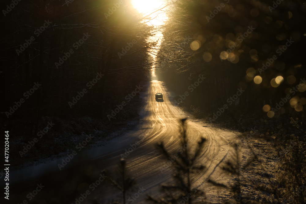 Sun rays through the trees and ice covered road with single car on it during the winter.