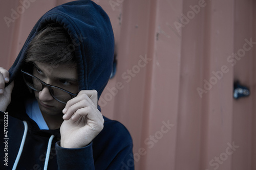 Mysterious teenager is hiding under a hood between garages. A thief and a burglar with glasses is hiding from the police. Fraud on the Internet. photo