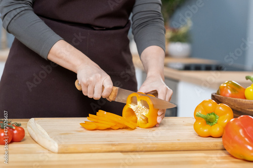 Hands of young female chopping fresh yellow capsicum on wooden board while going Fototapet