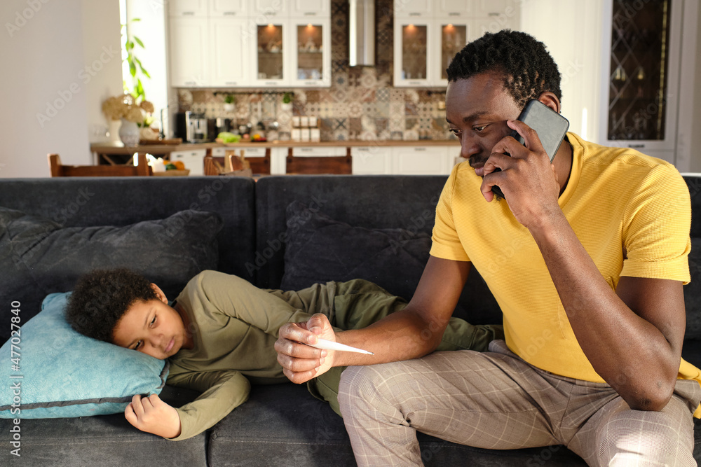 Young African man with smartphone by ear calling doctor for his sick little son while sitting on couch next to him