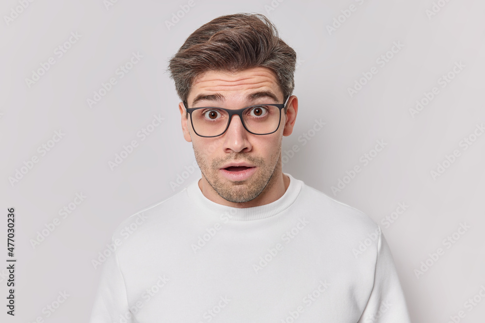 Shocked young man stares stupefied keeps mouth opened wears transparent glasses and jumper realizes something astonishing isolated over white background. Human reactions and emotions concept Shocked young man stares stupefied keeps mouth opened wears transparent glasses and jumper realizes something astonishing isolated over white background. Human reactions and emotions concept