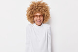 Photo of overjoyed curly haired European woman laughs out smiles toothily keeps eyes closed wears spectacles and turtleneck isolated over white background. Happy emotions and feelings concept