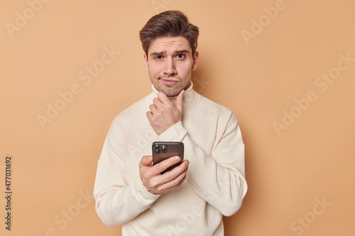 Puzzled thoughtful adult man holds chin and ponders on something holds mobile phone considers receieved notification thinks what answer to give wears turtleneck isolated on beige studio wall © wayhome.studio 