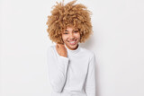 Portrait of lovely cheerful woman with curly bushy hair keeps hand on shoulder smiles pleasantly wears casual turtleneck enjoys good day has pleasant conversation with interlocutor poses indoor