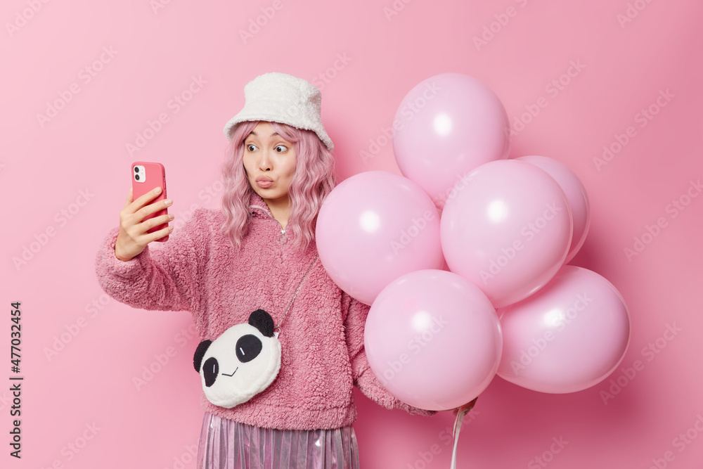 Lovely romantic Asian woman keeps lips rounded takes selfie via smartphone holds bunch of balloons going to congratulate friend with anniversary isolated over pink background. Celebration time Lovely romantic Asian woman keeps lips rounded takes selfie via smartphone holds bunch of balloons going to congratulate friend with anniversary isolated over pink background. Celebration time