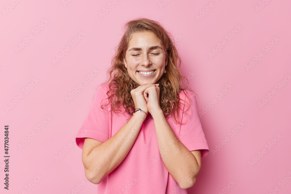 Happy cheerful European woman with dreamy expression has long wavy hair perfect teeth keeps hands under chin dressed in casual t shirt isolated over pink background. Positive emotions concept Happy cheerful European woman with dreamy expression has long wavy hair perfect teeth keeps hands under chin dressed in casual t shirt isolated over pink background. Positive emotions concept