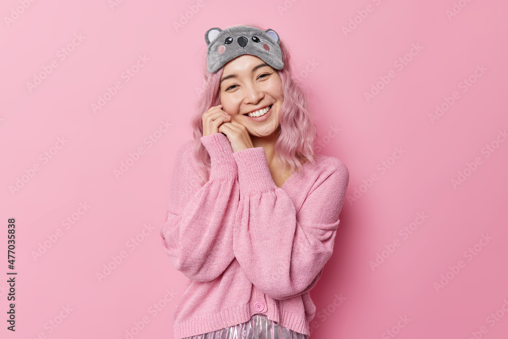 Pleased sincere woman keeps hands near face looks gently at camera going to go to bed after party wears jumper and sleepmask smiles positively isolated over pink background. Time for sleeping Pleased sincere woman keeps hands near face looks gently at camera going to go to bed after party wears jumper and sleepmask smiles positively isolated over pink background. Time for sleeping