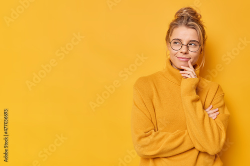 Pensive dreamy young European woman keeps hand near mouth looks suspicious away wears round spectacles and jumper isolated over yellow backgroud copy space aside for your promotional content photo