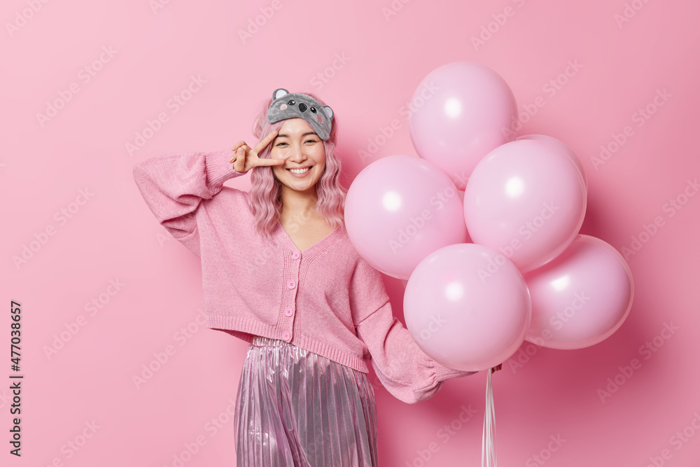 Positive good looking pink haired woman enjoys festive event comes to congratulate friend with birthday makes peace gesture over eye poses with helium balloons wears jumper skirt and sleepmask