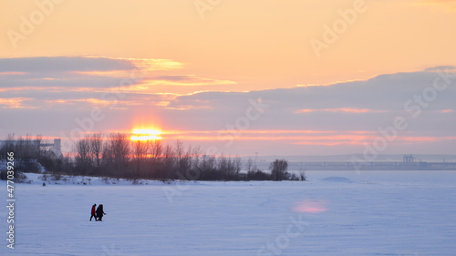  A group of young people crosses a frozen river on thin ice. It's getting late. The rays of the setting sun are reflected from the snow-covered ice surface. Copy space.