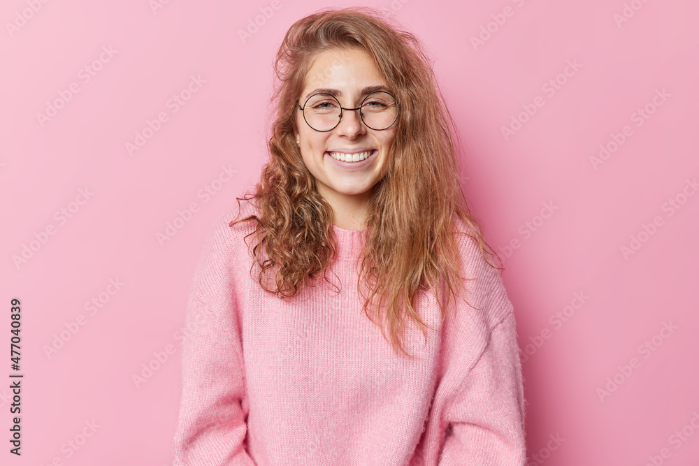 Positive young woman with gentle toothy smile feels glad wears round transparent glasses and sweater looks happily at camera isolated over pink background. Happy emotions and feelings concept Positive young woman with gentle toothy smile feels glad wears round transparent glasses and sweater looks happily at camera isolated over pink background. Happy emotions and feelings concept
