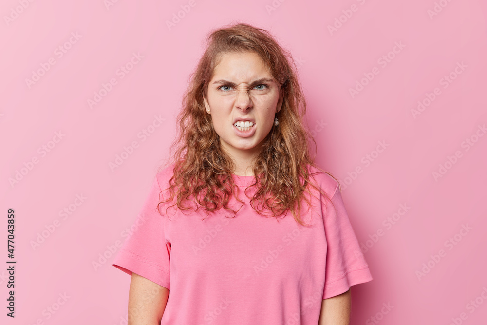 Portrait of angry irritated European woman smirks face feels outraged has annoyed expression clenches teeth wears casual t shirt isolated over pink background. Negative human emotions concept Portrait of angry irritated European woman smirks face feels outraged has annoyed expression clenches teeth wears casual t shirt isolated over pink background. Negative human emotions concept