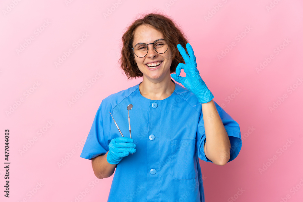 Woman English dentist holding tools over isolated on pink background showing ok sign with fingers Woman English dentist holding tools over isolated on pink background showing ok sign with fingers