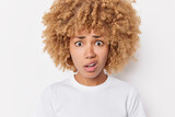 Headshot of displeased curly haired European woman purses lips with worried expression wears casual clothes poses against white background hears something unpleasant. Human reactions concept