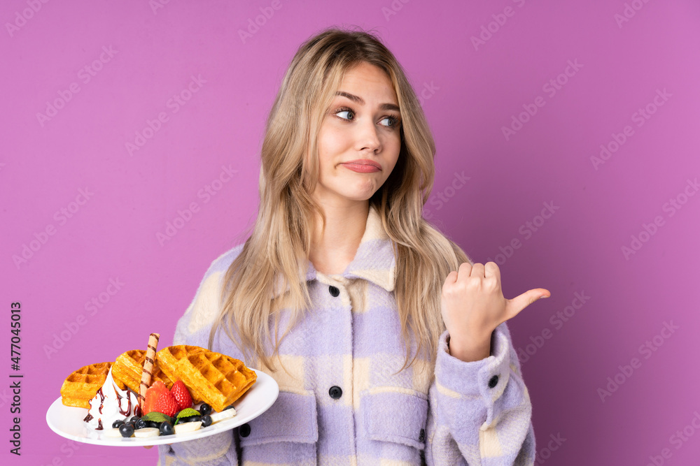 Teenager Russian girl holding waffles isolated on purple background unhappy and pointing to the side Teenager Russian girl holding waffles isolated on purple background unhappy and pointing to the side