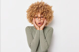 Photo of scared curly haired young woman grabs face looks with omg expression hears shocking news wears transparent glasses and poloneck isolated over white background. Human reactions concept
