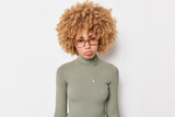 Upset woman with curly hair purses lips and looks sadly at camera has problems wears spectacles and poloneck isolated over white background. Offended female fed up of your excuses or justification