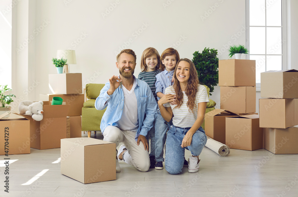 Happy family showing keys to their new home. Portrait of mom, dad and little children looking at camera and smiling in spacious living room full of cardboard boxes. Buying house or apartment concept Happy family showing keys to their new home. Portrait of mom, dad and little children looking at camera and smiling in spacious living room full of cardboard boxes. Buying house or apartment concept