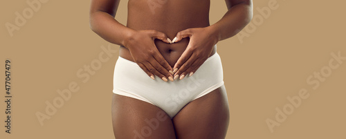 Foto Plus size black woman in underwear holding hands on abdominal area forming heart shape around navel, isolated on solid brown color background, cropped shot