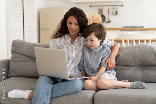Young mom teach preschool son to use laptop sit together with boy on couch in living room. Millennial single mother or babysitter have fun with child watch video or shopping online at home after work