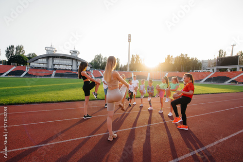 A large group of children, boys and girls, warm up and train under the guidance of a coach at the stadium during sunset. A healthy lifestyle.  © Andrii