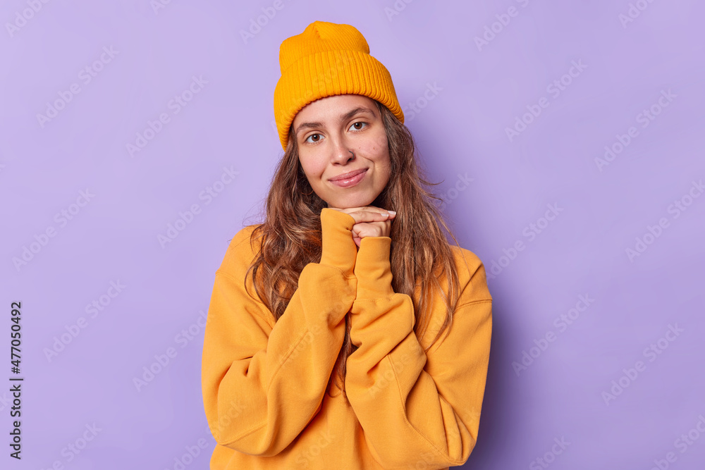 Charming long haired woman looks with tender expression keeps hands under chin wears casual orange jumper and hat isolated over purple background. People face expressions and felings concept Charming long haired woman looks with tender expression keeps hands under chin wears casual orange jumper and hat isolated over purple background. People face expressions and felings concept