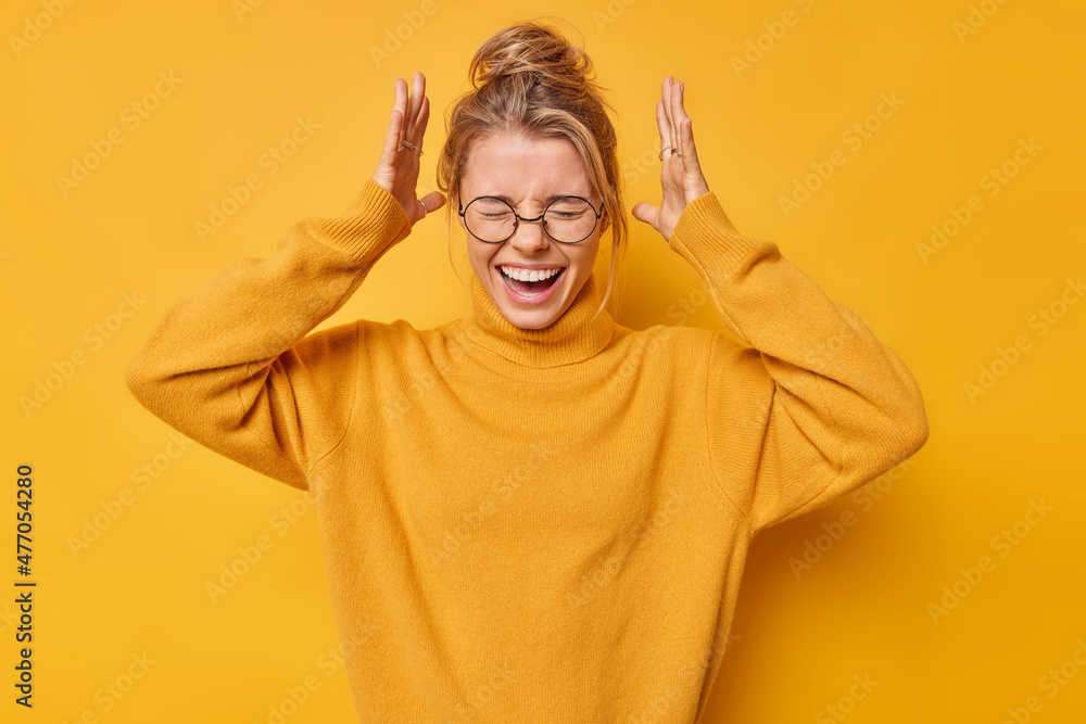 Emotional young European woman exclaims loudly keeps hands raised over head keeps eyes closed reacts on something awesome wears round spectacles casual jumper isolated over yellow background Emotional young European woman exclaims loudly keeps hands raised over head keeps eyes closed reacts on something awesome wears round spectacles casual jumper isolated over yellow background