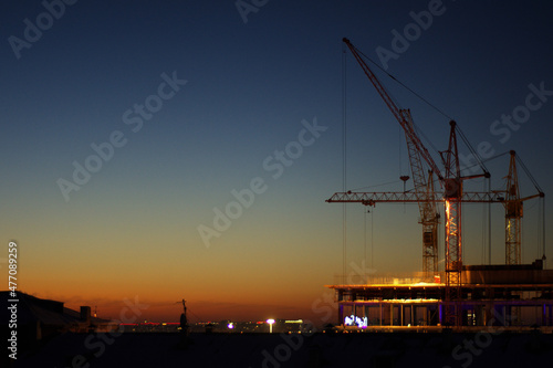 Construction cranes are building a house against the background of sunset and dark sky