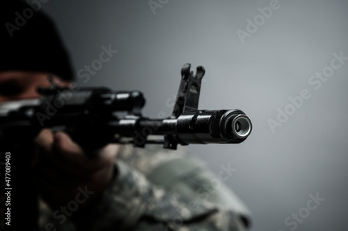 The muzzle of a Kalashnikov assault rifle. Close-up. A man in camouflage uniform and a black sniper hat takes aim before firing. Danger and military conflicts. Close-up. Gray background.