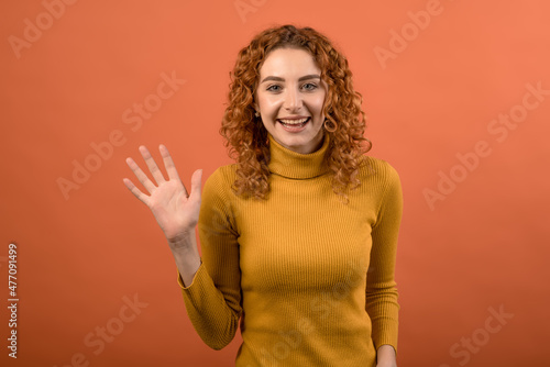 Young and attractive Caucasian redhead girl in orange jumper greeting and waving her hand isolated on orange studio background. © Павел Костенко