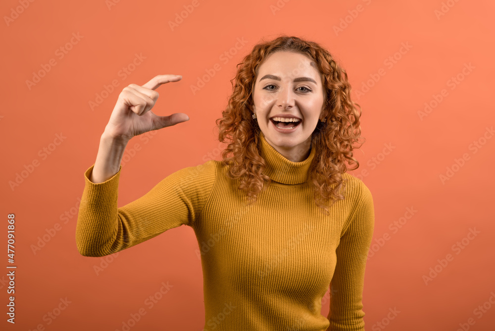 Young and attractive redhead Caucasian girl in orange jumper showing small size with her fingers isolated on orange studio background. Young and attractive redhead Caucasian girl in orange jumper showing small size with her fingers isolated on orange studio background.