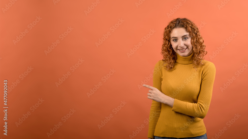 Young and attractive redhead Caucasian girl in orange jumper pointing her finger at copy space for advertising isolated on orange studio background. Young and attractive redhead Caucasian girl in orange jumper pointing her finger at copy space for advertising isolated on orange studio background.