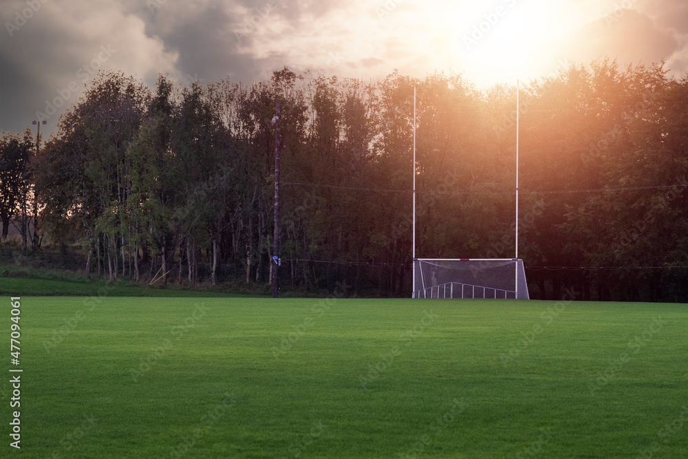 Tall goal post for Irish National sports on a training pitch against dark trees at sunset. Sun flare. Copy space. Camogie, hurling, rugby and Gaelic football playground. Calm mood. - obrazy, fototapety, plakaty 