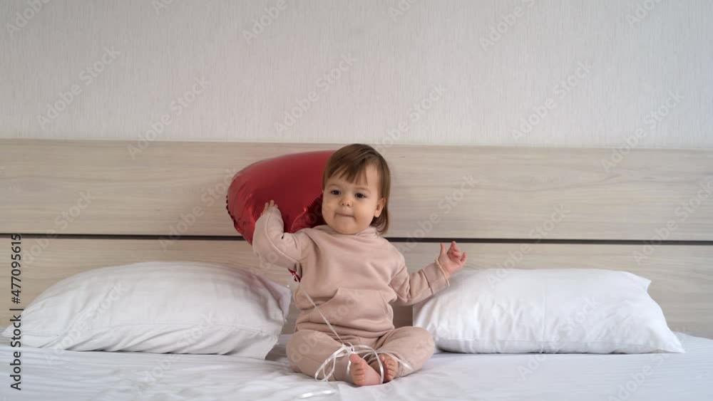 the child sits at home on the bed holding a red heart-shaped balloon on a rope, symbols of Valentine's Day. the child sits at home on the bed holding a red heart-shaped balloon on a rope, symbols of Valentine's Day.