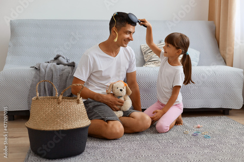 Horizontal shot of smiling young adult father spending time with his daughter while sitting on floor near sofa in living room, child with ponytails combing father's hair with colored tresses.