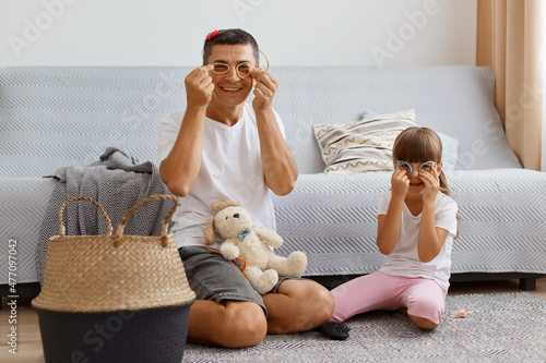 Image of dark haired male with his female child playing together at home, sitting on floor near cough in light room, making glasses with round scrunchies, having fun together, have funny expression.