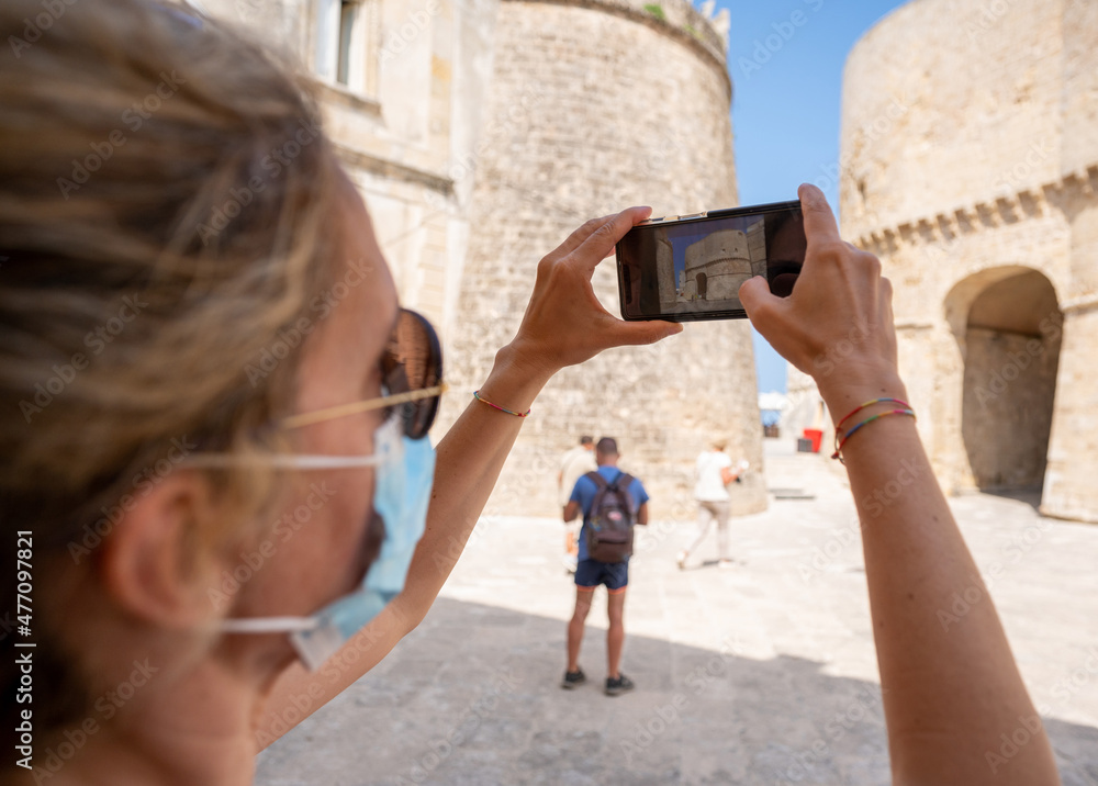 Otranto, Puglia, Italy. August 2021. A middle-aged woman with sunglasses and a coronavirus mask takes a picture of her with her smartphone inside the fort's walls. Otranto, Puglia, Italy. August 2021. A middle-aged woman with sunglasses and a coronavirus mask takes a picture of her with her smartphone inside the fort's walls.