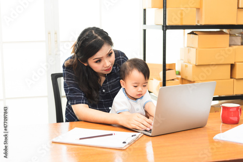 Beautiful Asian business mom is using a laptop while spending time with her cute baby boy at home for quarantine from virus crisis, work from home concept.